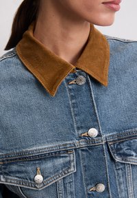 Veste en denim avec un col en velours côtelé marron, boutons en argent, deux poches poitrine et une texture bleue délavée.