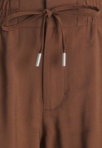 Brown fabric joggers with a smooth texture, featuring a drawstring waist and silver-tone tips, along with a side pocket detail.