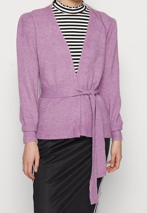 Femme portant un cardigan en tricot lavande avec une ceinture à la taille sur un haut à col montant rayé noir et blanc et une jupe noire.