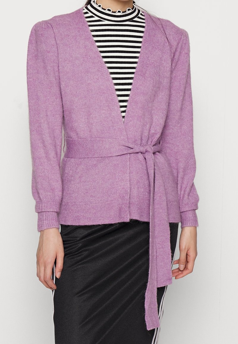 Femme portant un cardigan en tricot lavande avec une ceinture à la taille sur un haut à col montant rayé noir et blanc et une jupe noire.