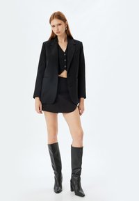 Schwarzes Blazer über einem schwarzen, cropped Top und einem Rock, kombiniert mit knielangen schwarzen Stiefeln. Glattes Material, taillierte Passform und minimalistisches Design.