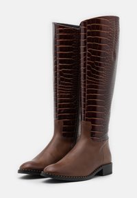 Bottes marron en cuir montant jusqu'au genou avec des panneaux à motif crocodile, talon bloc bas et surpiqûres blanches décoratives le long de la semelle.