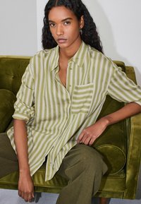 Camisa de botones a rayas verdes con bolsillo en el pecho, corte relajado, combinada con pantalones oliva. El fondo muestra un sofá de terciopelo verde.