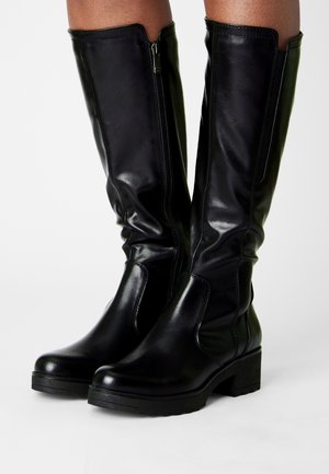 Botas de cuero negras hasta la rodilla con cremallera lateral y tacones gruesos, usadas por una persona que está de pie sobre una superficie blanca.