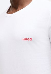 Balta medvilninė marškinėliai su apvalia apykakle ir raudonu "HUGO" logotipu kairiajame krūtinės pusėje. Lygus tekstūra ir klasikinis kirpimas.