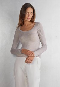 Helles graues Langarmoberteil aus einem weichen, strukturierten Stoff mit rundem Halsausschnitt und taillierter Silhouette. Kombiniert mit weißen Hosen.