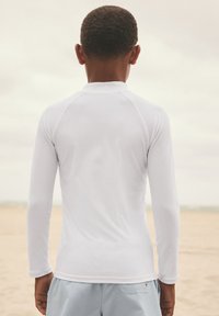 Maglia a maniche lunghe bianca con texture liscia, caratterizzata da una scollatura rotonda e cuciture piatte. Indossata con pantaloncini azzurri.