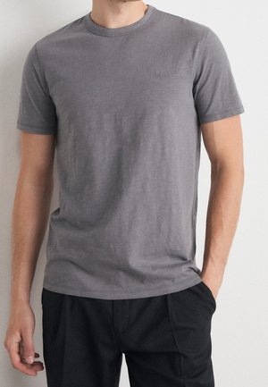 Person trägt ein eng anliegendes graues T-Shirt mit kurzen Ärmeln und dezent geprägtem "BOSS"-Logo sowie schwarze Hose, stehend vor weißem Hintergrund.