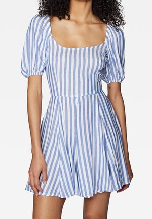 Femme portant une robe courte à rayures verticales bleues et blanches, avec des manches bouffantes et un décolleté carré, debout devant un fond uni.