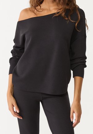 Vrouw draagt een zwarte off-shoulder top met lange mouwen en nauwsluitende zwarte broek, staat met ontspannen armen langs het lichaam.