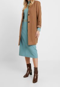 Brown wool coat, buttoned at front. Turquoise ribbed dress underneath. Brown patent leather ankle boots with square heels and crocodile pattern.