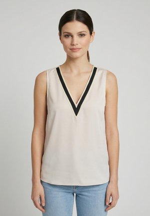 Mujer con cabello oscuro en una coleta baja, viste una camiseta sin mangas beige con un escote en V profundo y ribetes negros, combinada con vaqueros azul claro.