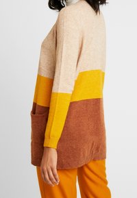 Vrouw die een kleurgeblokte trui met lange mouwen draagt in beige, geel en bruin, gecombineerd met oranje broeken, zijaanzicht ter hoogte van de taille.