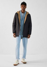 Chaqueta aislante negra y marrón, camisa de mezclilla por debajo, jeans azul claro, zapatillas blancas con suela beige, exhibidas en un modelo.