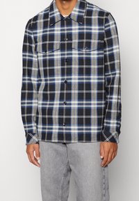 Koszula w kratę w kolorach granatowym, niebieskim i beżowym z dwoma kieszeniami na piersi, zapinana na guziki, z długimi rękawami, wykonana z teksturowanej tkaniny.
