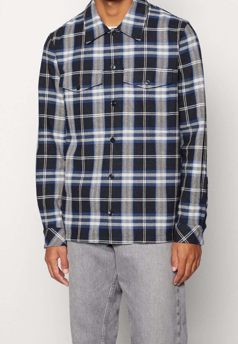 Koszula w kratę w kolorach granatowym, niebieskim i beżowym z dwoma kieszeniami na piersi, zapinana na guziki, z długimi rękawami, wykonana z teksturowanej tkaniny.