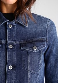 Närbild av en person som bär en blå denimjacka med silverknappar och en bröstficka, som visar detaljer på kragen och överkroppen.