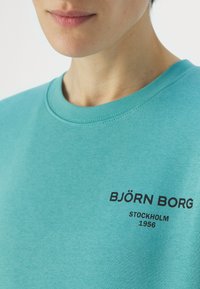 Sudadera turquesa hecha de tela suave. Cuenta con un cuello redondo y un texto de logo impreso en negro "BJÖRN BORG STOCKHOLM 1956" en el pecho.