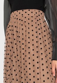 Jupe transparente à pois de couleur marron clair avec des pois noirs. Elle possède une taille haute et une texture fluide. Un haut noir contrastant est partiellement visible.