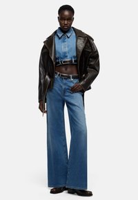 Brązowa skórzana oversize'owa kurtka, niebieska krótka jeansowa koszula i jeansy z wysokim stanem o rozszerzanych nogawkach, stylizowane z czarnym paskiem i brązowymi kozakami.