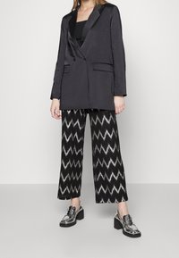 Czarna marynarka z satynowymi klapami, zestawiona z szerokimi spodniami w czarno-srebrny zygzakowy wzór oraz błyszczącymi srebrnymi butami na słupku.