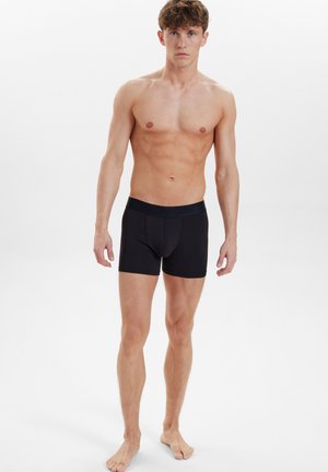 Schwarze Boxershorts mit glatter Textur, elastischem Bund und dezentem Branding, getragen von einem Model mit freiem Oberkörper.