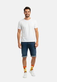 Camiseta blanca de manga corta, pantalones cortos de mezclilla azul, calcetines naranjas con patrón floral y zapatillas blancas. Atuendo sencillo y casual.