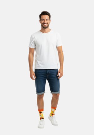 Weißes T-Shirt mit kurzen Ärmeln, blaue Jeansshorts, orangefarbene Socken mit Blumenmuster und weiße Sneakers. Einfaches und lässiges Outfit.