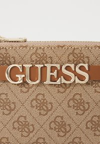 Brun mønstrete håndveske med en glatt tekstur, som har en fremtredende gull "GUESS"-logo og en kontrasterende brun lærrem.