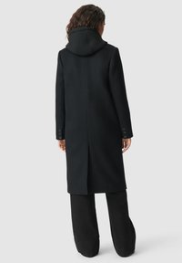 Vrouw in een lange zwarte jas met een capuchon, staande met haar rug naar de camera tegen een effen lichtgrijze achtergrond.
