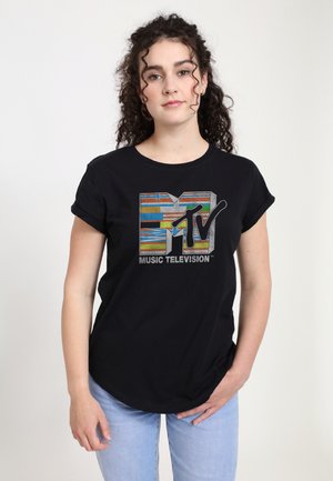 Femme aux cheveux bouclés portant un t-shirt noir MTV Music Television et un jean bleu clair, debout devant un fond uni.