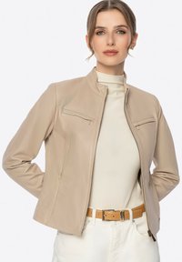 Veste en cuir beige avec un col montant, deux poches avant avec fermeture éclair, et une coupe ajustée. Portée par-dessus un haut clair et une ceinture.