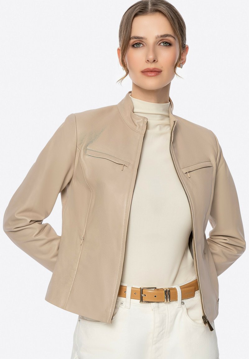 Veste en cuir beige avec un col montant, deux poches avant avec fermeture éclair, et une coupe ajustée. Portée par-dessus un haut clair et une ceinture.