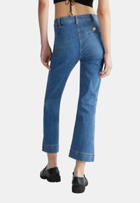 Jeans in denim di un blu medio con vita aderente e gambe svasate e accorciate. Presenta una tasca posteriore con un bottone decorativo. Texture liscia.