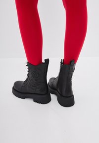 Cizme negre din piele până la gleznă, cu un model floral embosat, sistem de închidere cu șireturi în față, fermoar și talpă masivă, purtate cu colanți roșii.