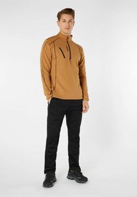 Brun zip-up fleece med sorte accenter, der har en høj krave og en frontlomme med lynlås, parret med sorte bukser og mørke fodtøj.