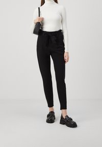 Pantalons noirs ajustés avec une taille à lien, associés à un col roulé côtelé blanc et des chaussures noires épaisses. Le look est complété par un petit sac noir.
