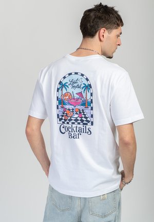 Mężczyzna zwrócony tyłem, ubrany w biały T-shirt z kolorową grafiką koktajlu i palmy oraz napisem „Lost Motel Cocktails Bar” na plecach.