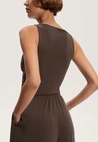 Dunkelbrauner ärmelloser Body mit Raffung an der Seite, elastischem Taillengummibund und seitlichen Taschen, der einen glatten, strukturierten Stoff präsentiert.