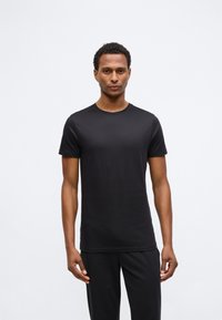 Tânăr stând cu fața spre înainte, îmbrăcat cu un tricou simplu negru cu mânecă scurtă și pantaloni negri, pe un fundal alb.