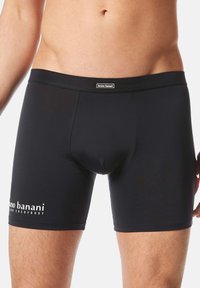 Męski tors w czarnych dopasowanych slipach z logo "bruno banani" na talii i lewym udzie, na jednolitym tle.
