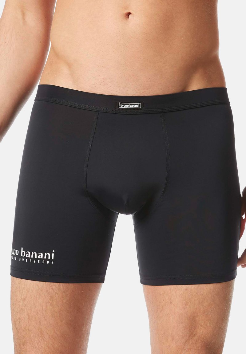 Męski tors w czarnych dopasowanych slipach z logo "bruno banani" na talii i lewym udzie, na jednolitym tle.