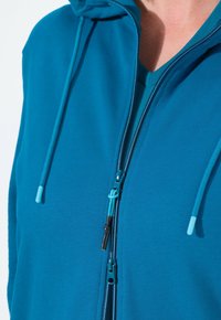 Teal zip-up huppari, jossa on säädettävä nauha, vetoketjullinen etuosa ja teksturoitu kangas. Huomionarvoisia mustia ja teal-värisiä varusteita.