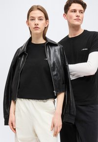 Νεαρή γυναίκα που φοράει μαύρο δερμάτινο μπουφάν πάνω από μαύρο πουκάμισο και ανοιχτόχρωμο παντελόνι, στέκεται δίπλα σε νεαρό άνδρα με στρωτά μαύρα και λευκά ρούχα.