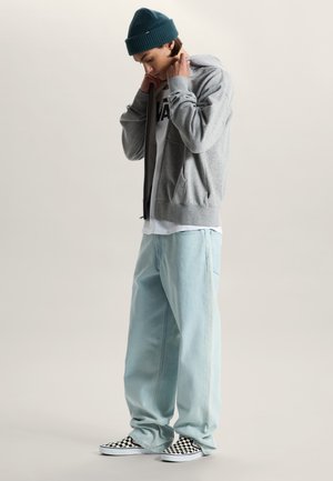 Jongere draagt een groene muts, grijze hoodie, wit Vans T-shirt, lichtblauwe losse spijkerbroek en zwart-wit geruite instap-schoenen.