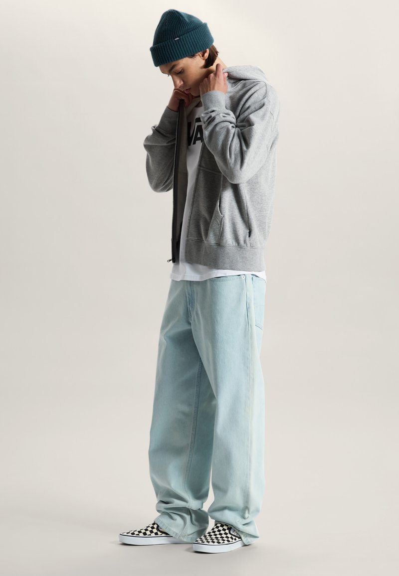 Joven con un gorro de color verde azulado, sudadera con capucha gris, camiseta blanca de Vans, vaqueros anchos azul claro y zapatos slip-on a cuadros en blanco y negro.