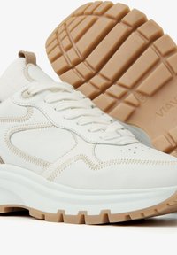 White sneaker with leather and textile materials, featuring a thick white sole and a textured tan outsole. Stitching and perforations are visible.