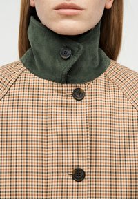 Personne portant un manteau à carreaux marron et beige avec un col montant en velours côtelé vert fixé par un bouton noir.