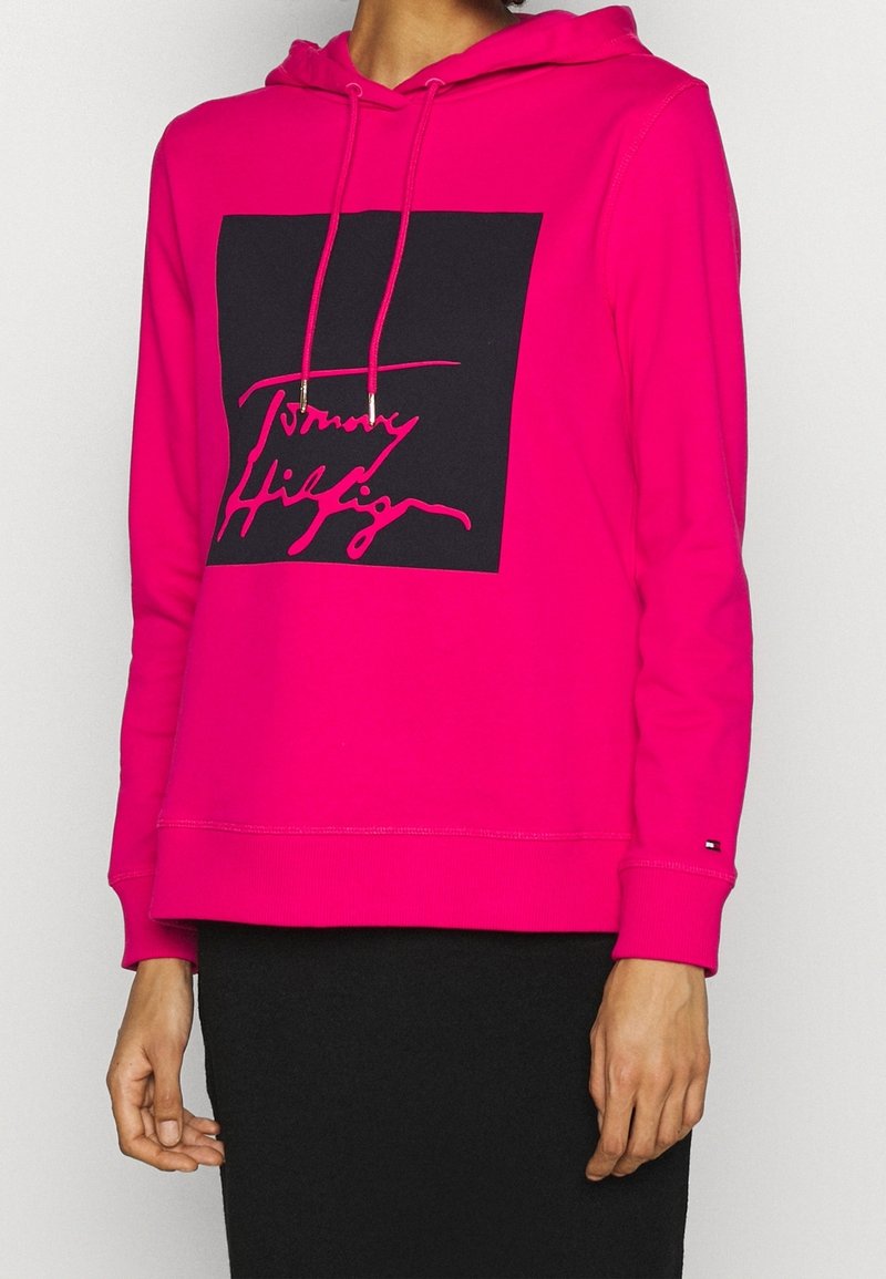 Sweat à capuche rose vif avec cordons, arborant un grand carré noir et un logo en cursive rose sur la poitrine, porté avec une jupe noire.