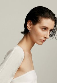 Una persona con pelo corto y con aspecto mojado lleva una blusa blanca sin hombros. Tienen pequeños pendientes colgantes con un diseño distintivo.